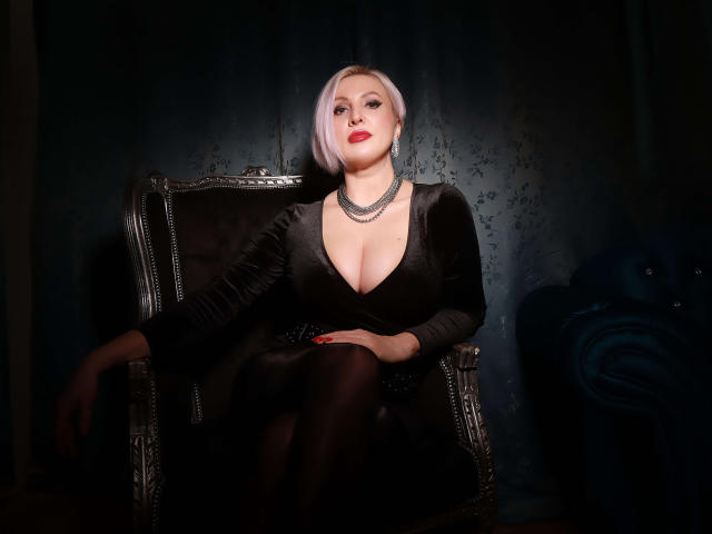NicoleDomme sits in a dark, velvet chair, wearing a black dress with a deep cleavage, showing her tits, and black tights, with a short, white bob and red lipstick.