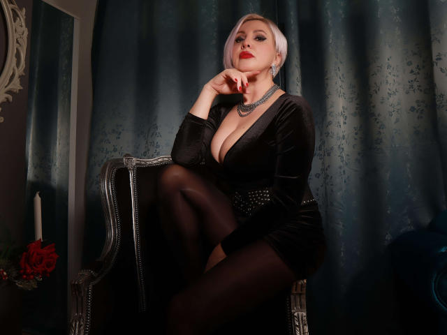 NicoleDomme with pale skin, white hair, red lipstick, black velvet dress, deep cleavage, black stockings, sitting on ornate chair, hand on chin, looking seductive.