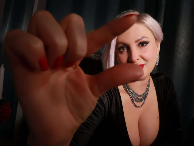 NicoleDomme with pale pink hair, red lipstick, black dress, and silver necklace, pointing at the camera with red-painted fingers, showing off her big boobs in a dark room.