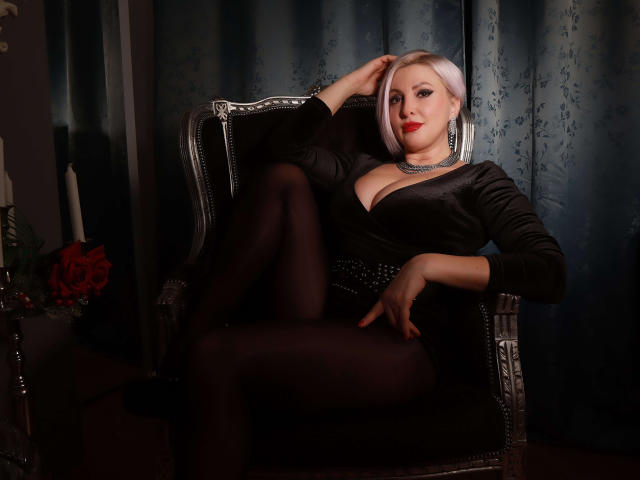 NicoleDomme with blonde bob, red lipstick, black dress, and black tights, sitting in a silver chair, hand on head, legs crossed, showing her big boobs and ass.
