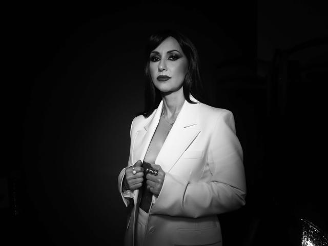 Black-and-white photo of MissVonTease with dark hair, wearing a white blazer, exposing her tits, holding a microphone, looking confident, with makeup and dark lipstick.