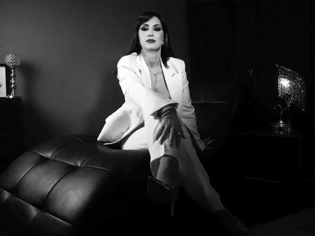 MissVonTease, com cabelo escuro, vestindo um terno branco, sentada em um banquinho de couro preto, com a mão no joelho, com uma expressão confiante, saltos pretos, batom escuro, foto em preto e branco..