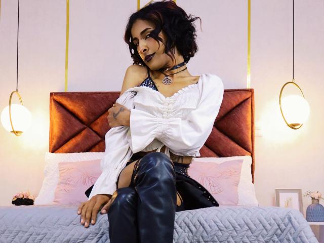 NoemiSky with dark makeup, black choker, white off-shoulder shirt, black leather pants, and thigh-high boots, sitting on a bed with a red velvet headboard, arms crossed.