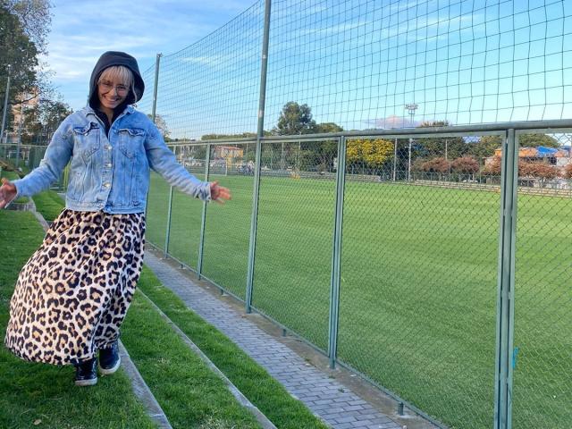 SaraleePoison with a blue denim jacket and leopard print skirt, smiling, arms out, standing in front of a green fence and grassy field. She's wearing black shoes.