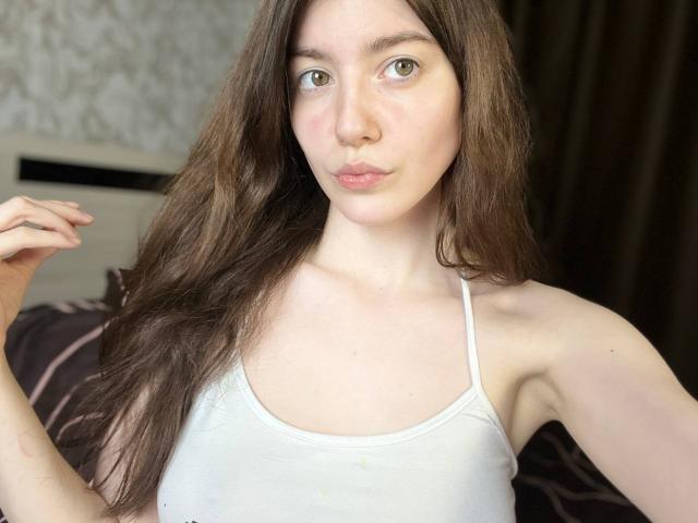 VinnaSkyHott mit langen, braunen Haaren, die ein weißes Tanktop trägt, wobei ihre Brüste sichtbar sind, mit sinnlich geschürzten, rosafarbenen Lippen, heller Haut und grünen Augen, in einem leicht unscharfen Selfie..