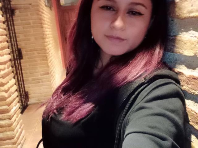 LiaJonnesX with purple-tipped hair, black top, and subtle eyeliner, smiling, holding a black bag, standing against a stone wall. Boobs visible, black zipped jacket.