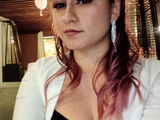 LiaJonnesX with pink-dyed hair, wearing a white jacket and black top, showing her tits, has a serious expression, wearing sparkly earrings, in a dimly-lit room.