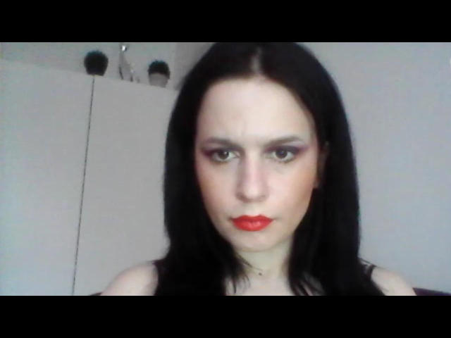 ScarlettAmore, con piel pálida, cabello negro, lápiz labial rojo y sombra de ojos rosa, con una expresión seria, vistiendo una blusa negra, mostrando ligeramente sus senos..