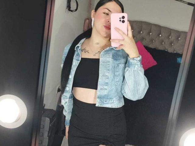EmmieX with black hair, wearing a denim jacket, black tube top, and black ribbed skirt, holding a pink phone, showing tattoos on her chest, mirror selfie.