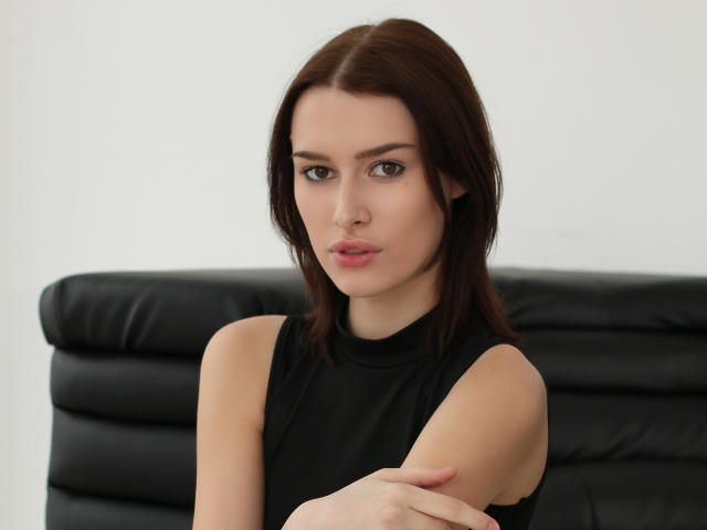 AmandaGate with brown hair, black sleeveless top, pale skin, and pink lips, arms crossed, looking straight at camera, seated on black leather couch.
