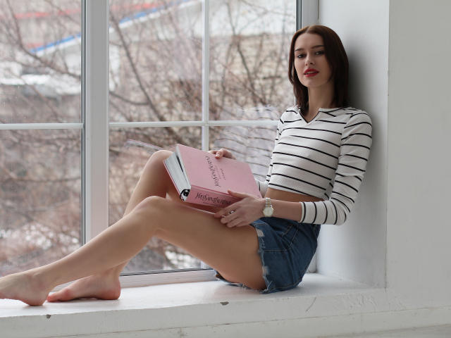 AmandaGate with tits, wearing a striped shirt and denim shorts, sits on a window ledge, reading a pink book, legs crossed, barefoot, with a wristwatch, looking at camera.
