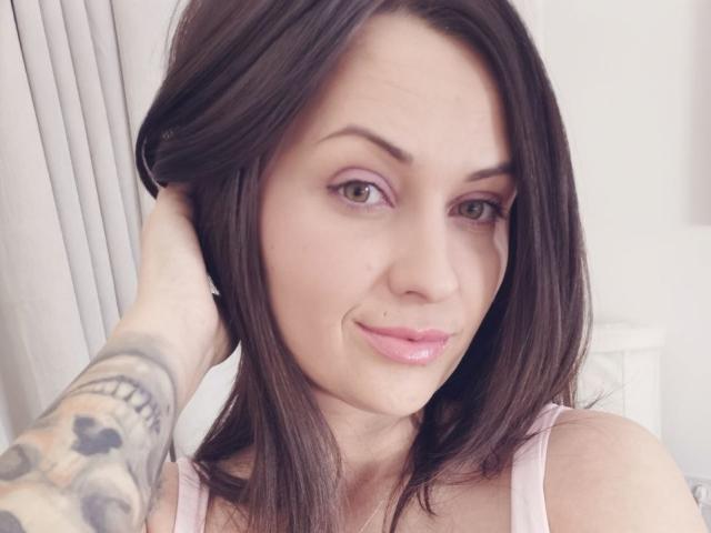 Cali69 tiene cabello castaño, ojos verdes, lápiz labial rosa, un piercing en la nariz y tatuajes en la mano derecha, sonriendo con una mano en el cabello, y lleva una camiseta sin mangas rosa..