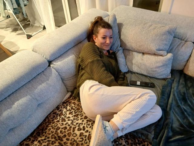 CoupleRealHot with brown hair in a ponytail, green sweater, white ribbed pants, and blue sneakers, lying on a leopard print rug, remote control near her ass.