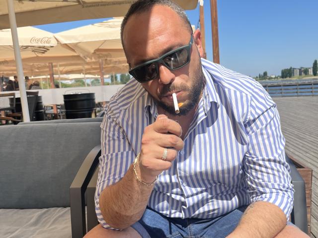 CarlitoBrigante, wearing striped shirt and sunglasses, holding cigarette, with butt in mouth, has ring on finger, sitting on gray chair, looking down, blue sky in background.