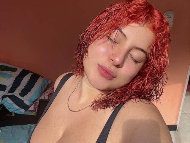 RoseDijon with red, wet, curly hair, eyes closed, smiling softly, wearing a black tank top, showing her big, round boobs, fair skin, and a simple necklace.