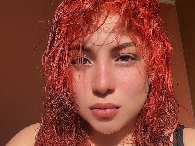 RoseDijon has messy red hair, fair skin, and is looking at the camera with a slight pout, wearing a black tank top, showing her bare shoulders.