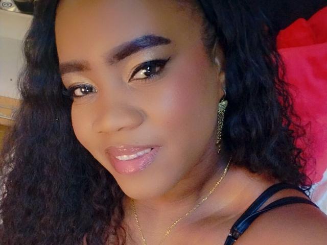 DanielaBrown's face with dark brown skin, curly black hair, and glossy pink lips. She's wearing black eyeliner, gold earrings, and a gold necklace, looking slightly to the side.