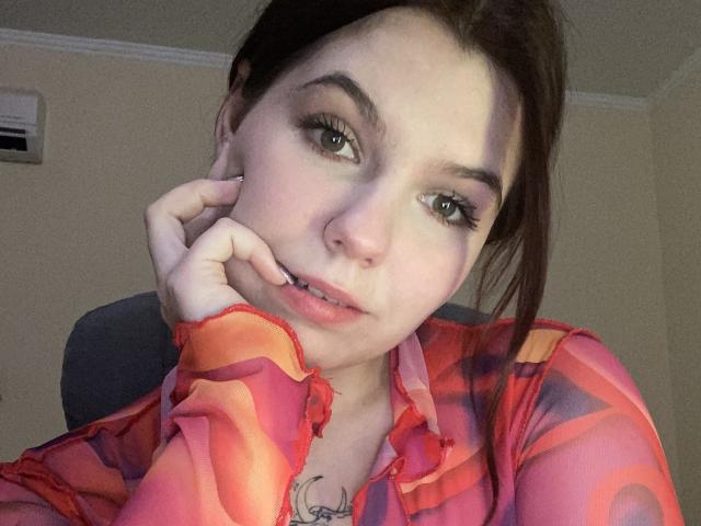 Khaleessi with brown hair, green eyes, and light skin, wearing a red and orange patterned shirt, resting her chin on her hand, showing a small tattoo on her neck.