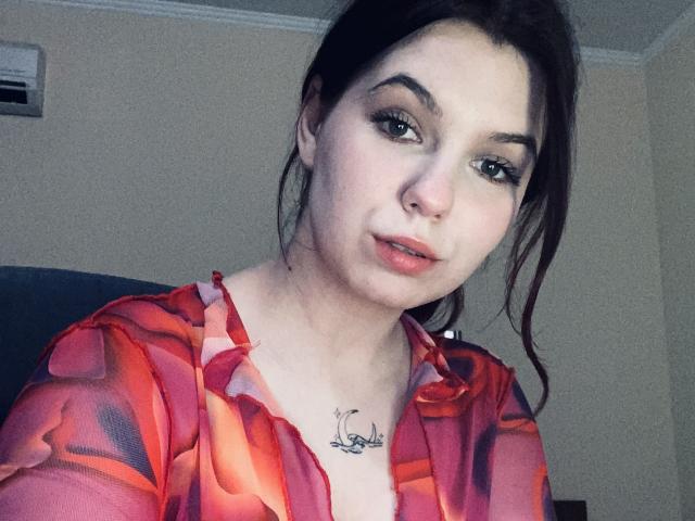 Khaleessi with pale skin, dark brown hair in a ponytail, red-orange sheer top, green eyes, and a small nose ring, showing her tits and a cursive tattoo below.