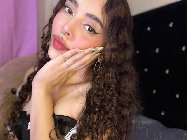 TianaVera with curly brown hair, nose ring, and green nails, touching her cheek, wearing black lace, showing her tits, and a ring on her finger.