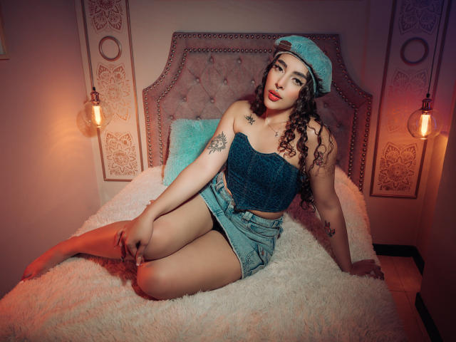 TianaVera with curly dark hair, wearing a blue cap, textured blue crop top, and denim shorts, sitting on a fluffy bed, showing tattoos and a sexy, confident look.