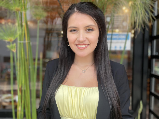 AmelieMuller has long black hair, a cute smile, and is wearing a yellow top with a black cardigan, and a small butterfly necklace. Her tits are covered by her top.