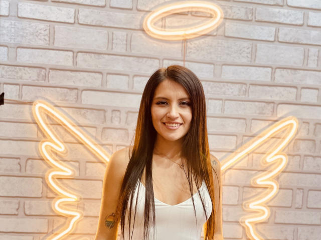 AmelieMuller, with long brown hair and white tank top, smiling, has small tits and a tattoo on her left arm, standing in front of neon angel wings and halo.