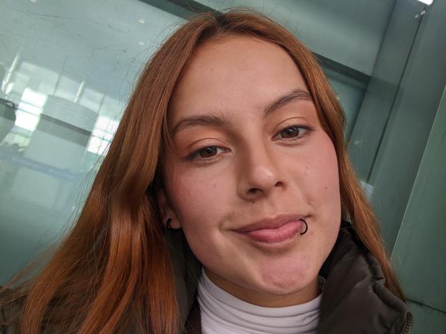 VictoriaOrtega has light brown hair, a slight pout with tongue out, and is wearing a green jacket over a white turtleneck. Her skin is fair with visible pores.