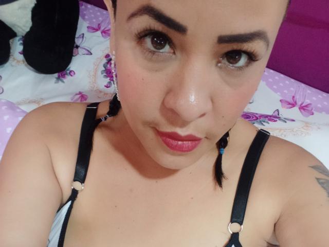 MimiP with shiny face, brown eyes, and pink lipstick, wearing black straps, looking up with a slight pout, her boobs partially visible, short black hair, tattoo on right shoulder.