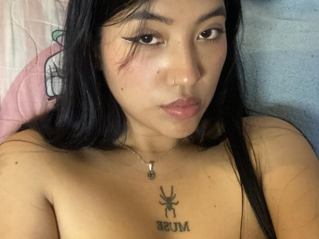 Elizaa tem cabelos longos e pretos, delineador de olhos estilo "gatinho" e uma tatuagem de abelha com a palavra "BZUM" abaixo, no peito. Ela está sem blusa, com um pequeno piercing no nariz e um colar..