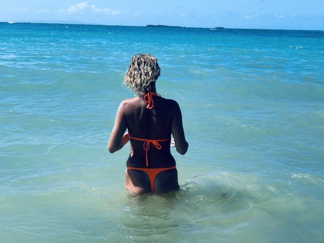 BlackAngeel with wet, curly blonde hair, wearing an orange bikini, stands in clear blue water, back to camera, showing her tan skin and small orange bikini bottom.