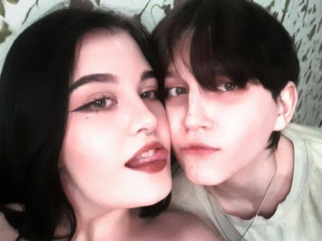 RavenRadiant with dark hair and eyeliner, nose ring, and red lipstick, next to a pale, brown-haired person with earbuds, both looking at the camera.