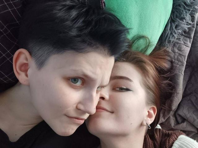 RavenRadiant with short black hair and pale skin is lying next to a red-haired RavenRadiant with eyeliner, both with subtle smiles, close faces, and brown pillows.