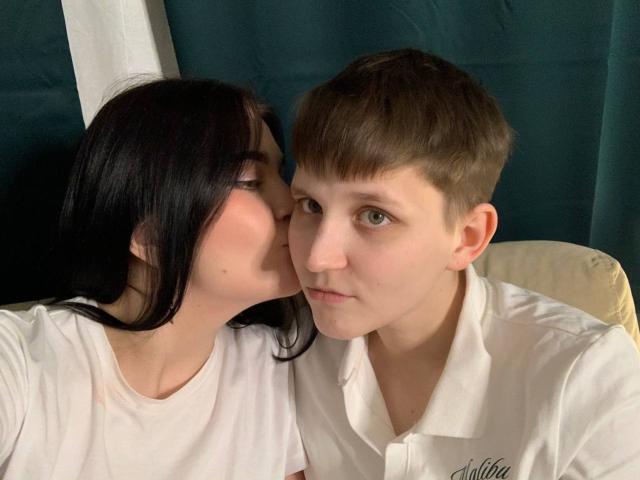 RavenRadiant with black hair kissing the cheek of a short-haired boy in a white shirt, both wearing white shirts, looking into the camera. His face is slightly flushed.