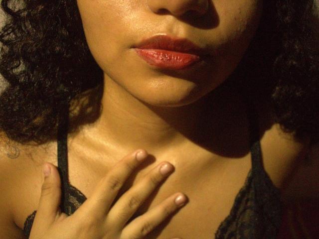 SexLaddy's face, focusing on her glossy red lips and dark curly hair, with a hand touching her chest, wearing a black lace top, in warm, dim lighting.