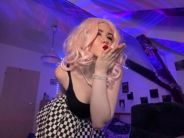 EvaxFoxy with pink curly hair, black top, checkered skirt, red lipstick, kissing fingers, wearing pearl bracelet, in dimly lit room with blue-purple lights, showing her tits.