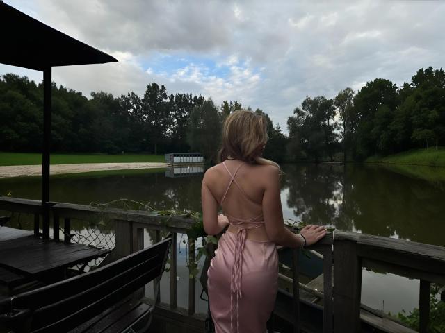 EvaxFoxy with blonde hair, wearing a pink, backless dress showing her ass, standing on a wooden deck, facing a calm, reflective pond, with trees and a cloudy sky.
