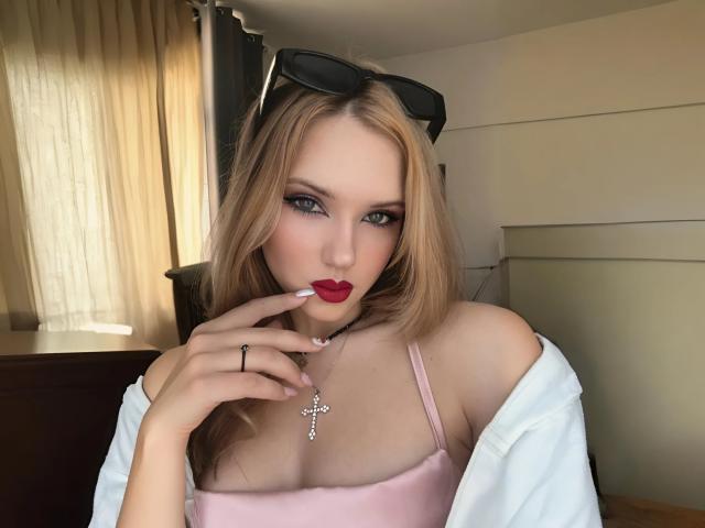 EvaxFoxy with blonde hair, red lipstick, pink top, white shirt off shoulders, black sunglasses on head, finger near mouth, cross necklace, black ring, light skin, blue eyes.