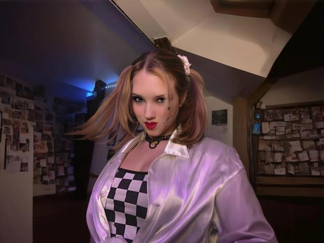 EvaxFoxy with puffy pink-tinted hair in pigtails, wearing a shiny purple jacket, black-and-white checkered top, and black choker, standing in dimly lit room with red carpet.