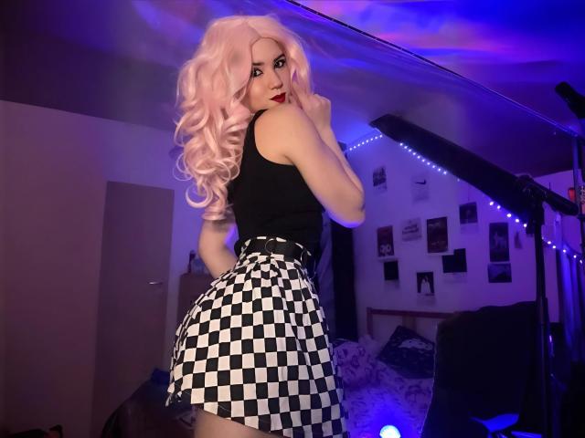 EvaxFoxy with pink curly hair, black tank top, and black-and-white checkered skirt, looking back, showing her ass, in a dimly lit room with blue lighting.
