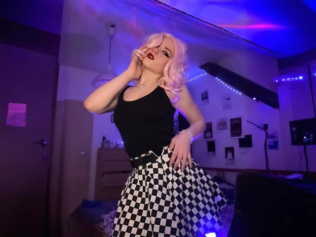 EvaxFoxy with pink hair, black tank top, and checkered skirt, biting red lipstick, hand on hip, in dimly lit room with purple and blue lights.