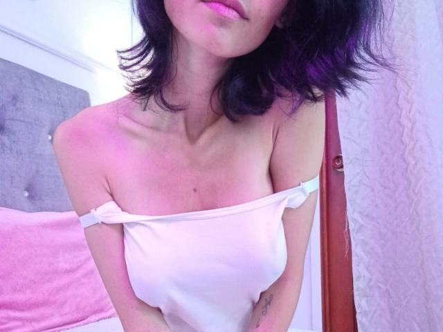 Jasmin Garcia, con una maglietta bianca senza spalline che mette in mostra piccoli seni, rossetto rosa, capelli scuri e ondulati, un tatuaggio sul braccio destro, in piedi in una camera da letto con un letto rosa e una parete bianca..