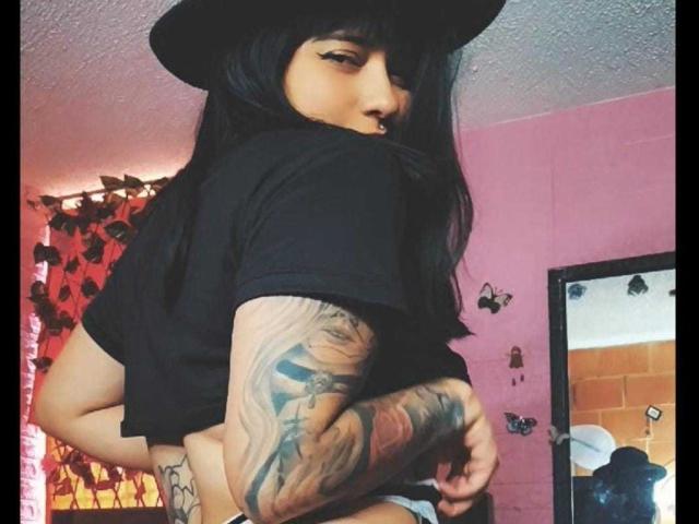 Scarletdeepthoat69 con sombrero negro, cabello negro largo, camisa negra, brazo tatuado y piercing en la nariz, mirando hacia atrás, mostrando el trasero desnudo. Habitación con paredes rosas y espejo..
