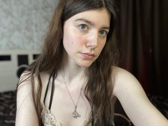 VinnaSkyHott with long brown hair, green eyes, and a small mole on her left cheek, wearing a lace top, necklace with a cross, and looking at the camera.