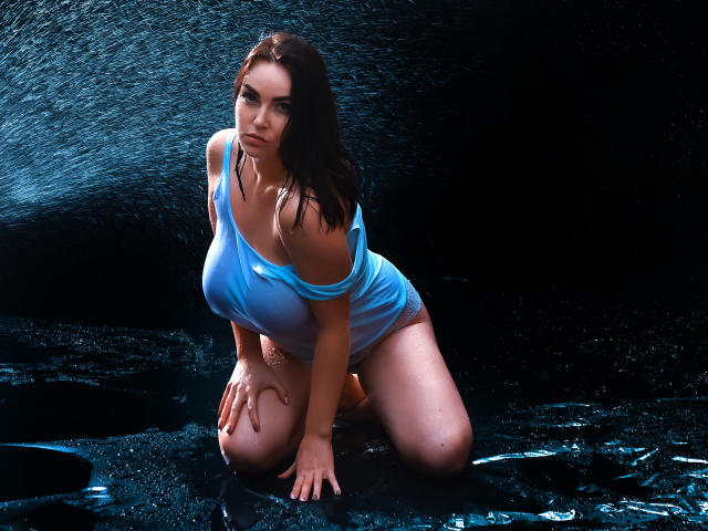 WilsonMelanie kneeling in water, wearing a wet, see-through blue shirt, exposing her large tits and bare ass, with dark wet hair and a serious expression.