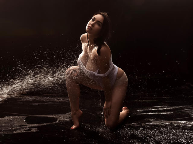 WilsonMelanie kneels on wet black ground, wearing a white, wet, see-through shirt, water splashing, exposing her tits, with a dark, moody background. Her head is tilted back.