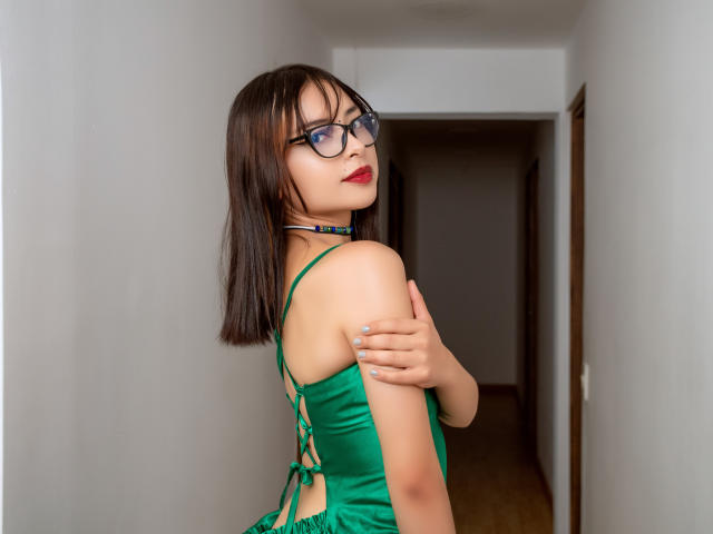 EvyEmber con un vestido verde, gafas y lápiz labial rojo, con los brazos cruzados, mostrando sus senos, con un corte de pelo bob castaño, mirando por encima del hombro, en un pasillo..