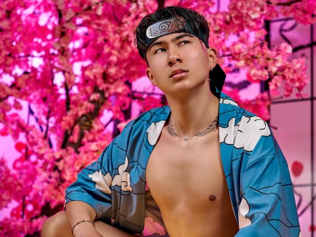 NoahAndRose, con un pecho delgado y tonificado, vistiendo un kimono floral azul, una diadema negra con un símbolo ninja y un tatuaje debajo del mentón. Flores de cerezo rosadas en el fondo..