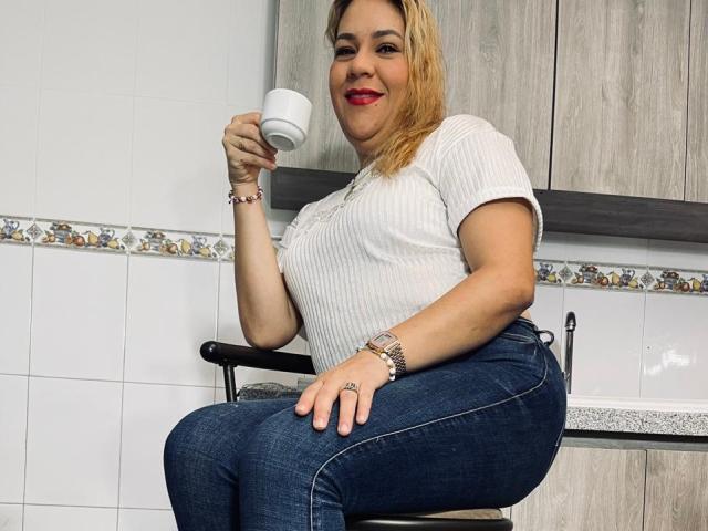 Mya Lopez, con i capelli biondi, rossetto rosso, maglietta bianca a coste, jeans blu, che tiene una tazza bianca, seduta su uno sgabello da cucina, sorridente, che indossa un braccialetto e un orologio, mostrando il sedere..