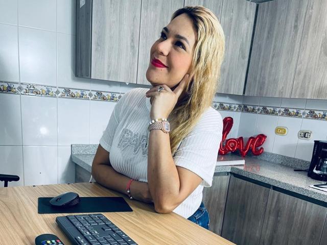 MyaLopez with blonde hair, red lipstick, white ribbed shirt, resting chin on hand, smiling, sitting at kitchen table, wooden countertop, gray cabinets, red "love" sign, keyboard in front.